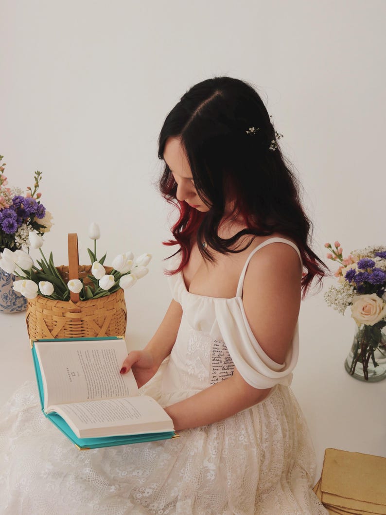 K&ouml;nnte beinhalten: Eine Frau in einem wei&szlig;en Spitzenkleid mit einem Blumenhaarband liest ein Buch mit t&uuml;rkisfarbenem Einband. Sie sitzt auf einem Stapel B&uuml;cher mit Blumen in Vasen auf beiden Seiten von ihr.