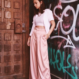 May include: A woman wearing a white crop top, a white button-down shirt, and pink wide-leg pants. She is standing in front of a wooden door and a wall with graffiti.