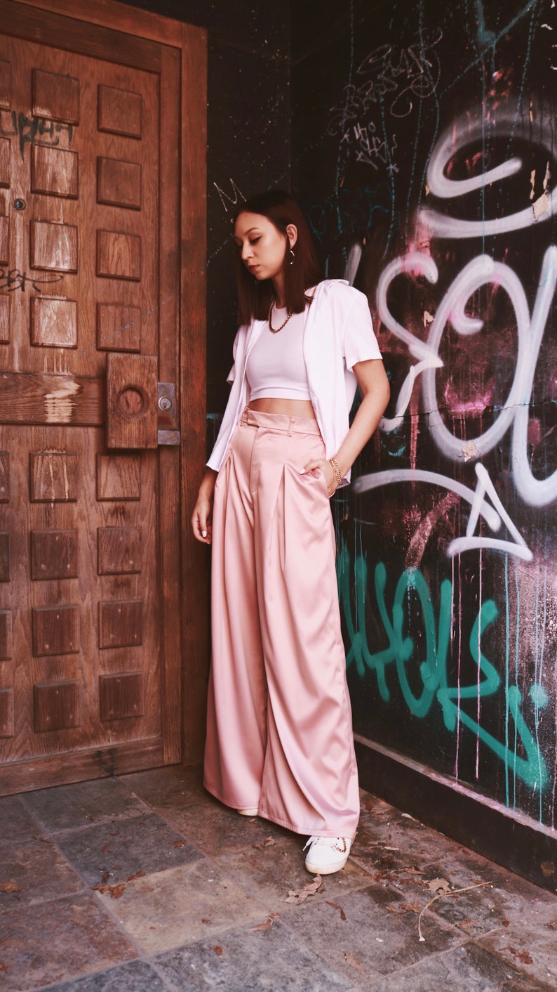 May include: A woman wearing a white crop top, a white button-down shirt, and pink wide-leg pants. She is standing in front of a wooden door and a wall with graffiti.
