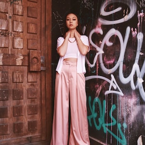 May include: A woman wearing a white crop top and a white cardigan with a pink pair of wide-leg pants. She is standing in front of a wall with graffiti.
