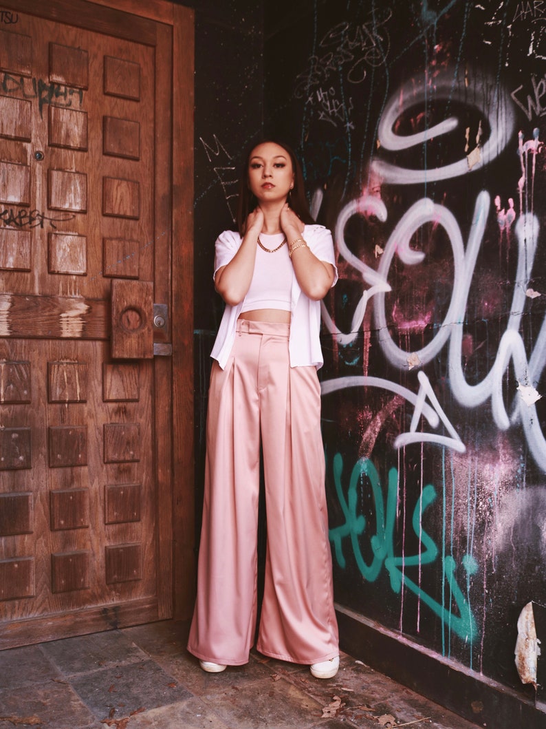 May include: A woman wearing a white crop top and a white cardigan with a pink pair of wide-leg pants. She is standing in front of a wall with graffiti.