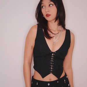 May include: A woman wearing a black corset top with lace-up detailing and a black skirt with silver embellishments. She is wearing multiple silver necklaces with a padlock charm.