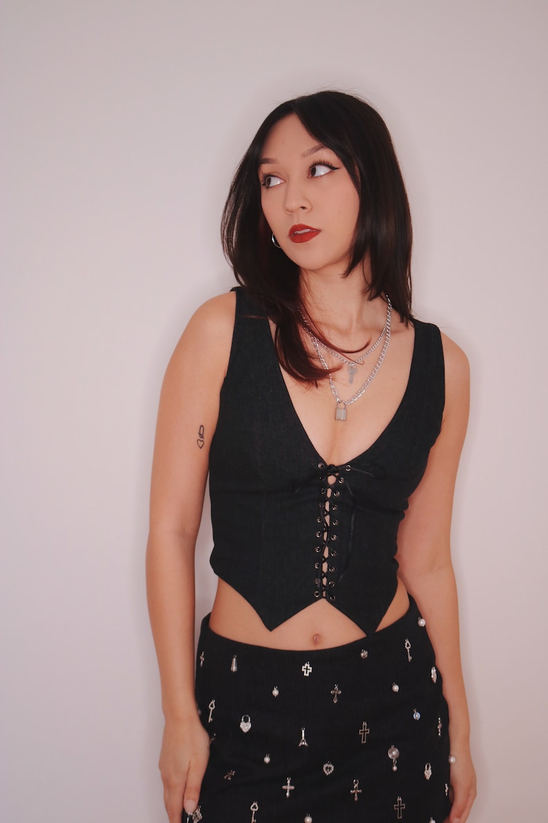 May include: A woman wearing a black corset top with lace-up detailing and a black skirt with silver embellishments. She is wearing multiple silver necklaces with a padlock charm.