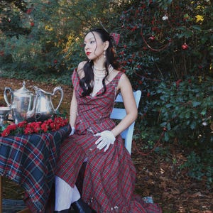 May include: A woman in a red plaid dress sits at a table set for tea. The dress has a unique design with a draped neckline and suspender straps. The table is covered with a matching plaid tablecloth and has a silver tea set and red flowers.