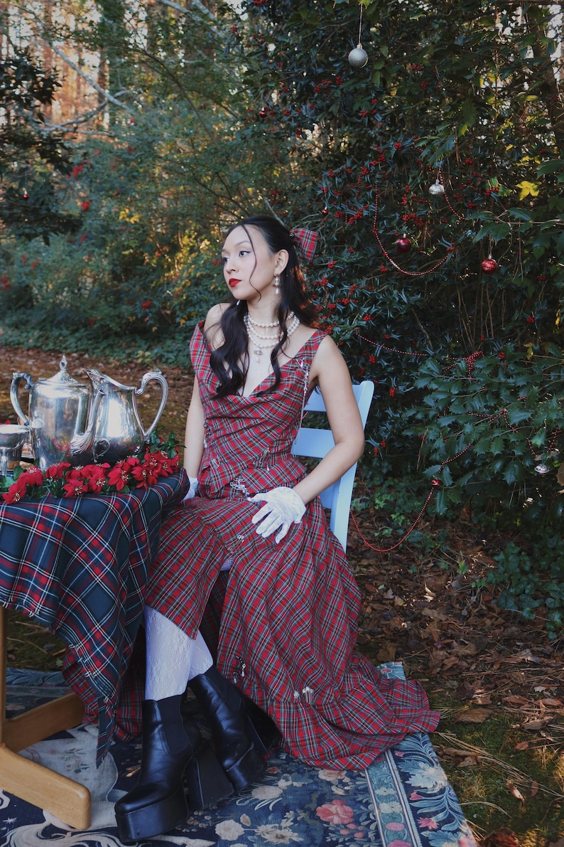 May include: A woman in a red plaid dress sits at a table set for tea. The dress has a unique design with a draped neckline and suspender straps. The table is covered with a matching plaid tablecloth and has a silver tea set and red flowers.