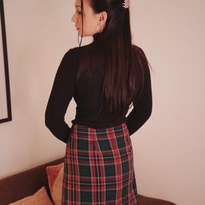 May include: A black long-sleeved turtleneck top paired with a plaid mini skirt in red, green, and white. The outfit is completed with black tights. A hair clip is visible in the hair.