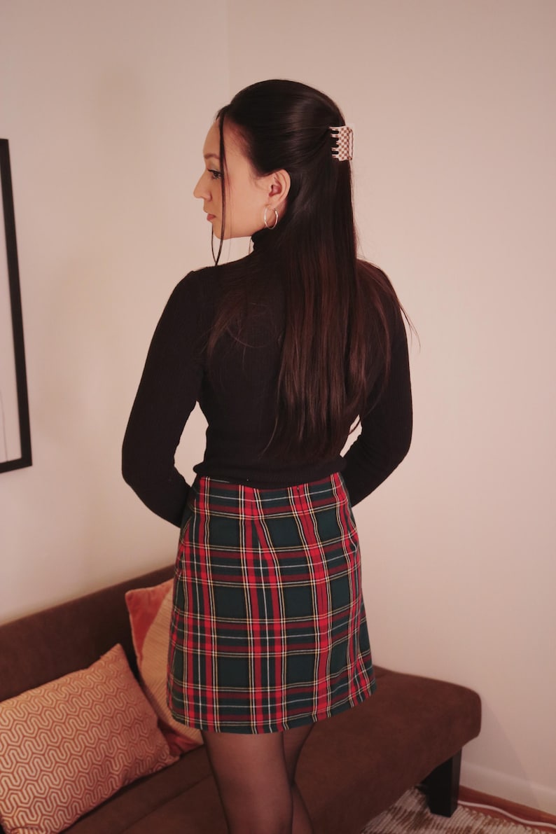 May include: A black long-sleeved turtleneck top paired with a plaid mini skirt in red, green, and white. The outfit is completed with black tights. A hair clip is visible in the hair.
