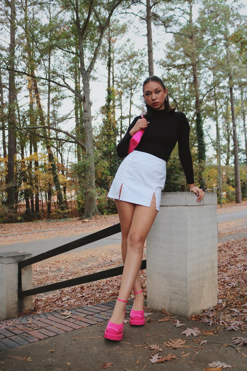 May include: A woman wearing a black turtleneck shirt, a white mini skirt with a side slit, and pink platform heels. She is holding a pink handbag and standing in a wooded area.