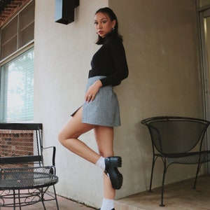 May include: A woman wearing a black turtleneck sweater and a plaid skirt stands with one leg bent, wearing black platform shoes and white socks. She is standing in front of a white wall with a black rectangular light fixture.