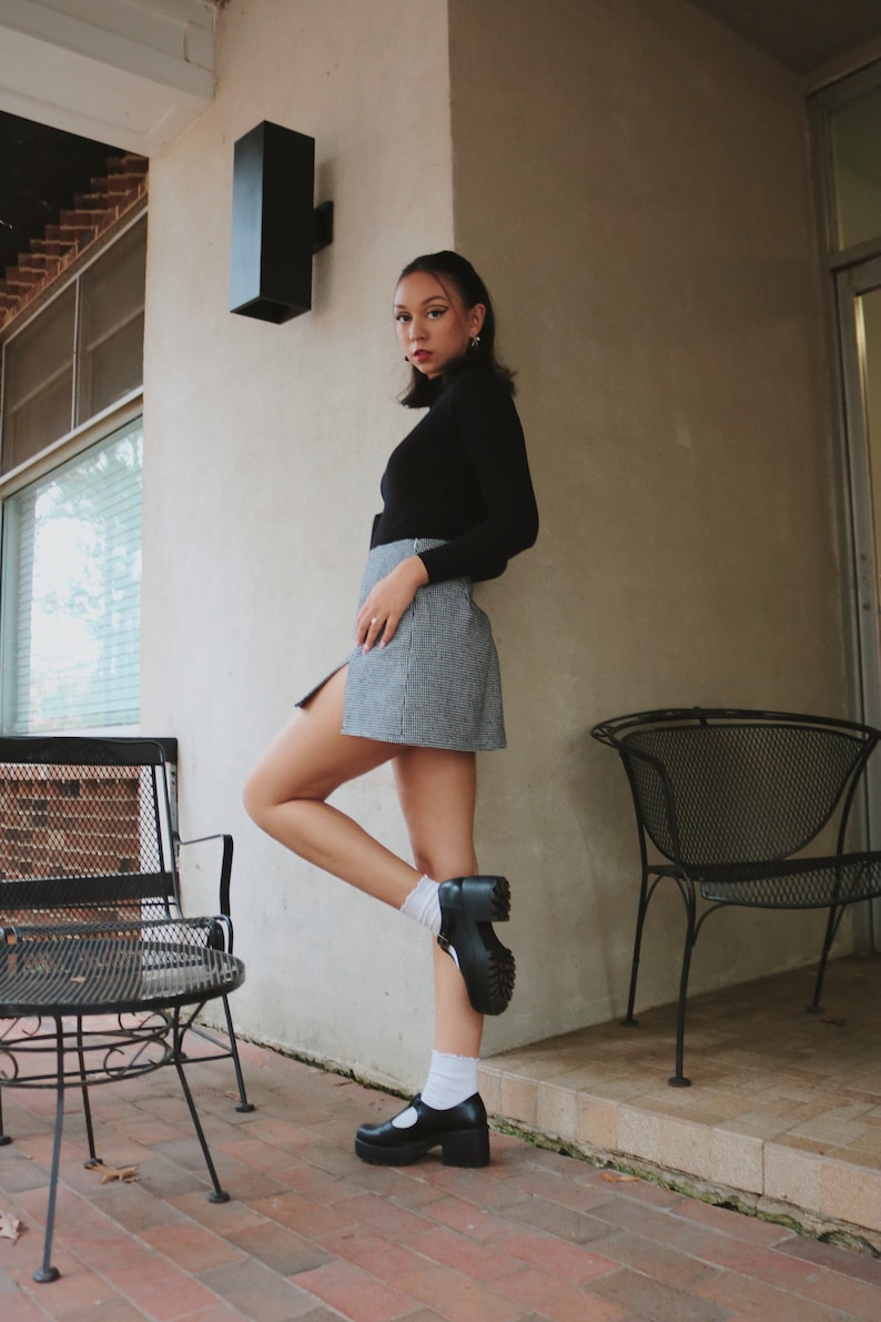 May include: A woman wearing a black turtleneck sweater and a plaid skirt stands with one leg bent, wearing black platform shoes and white socks. She is standing in front of a white wall with a black rectangular light fixture.