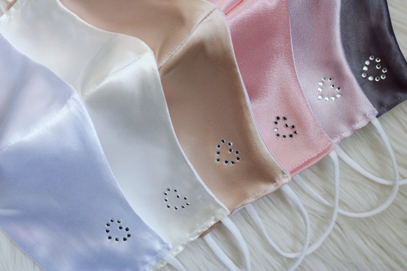 May include: Five satin face masks in different colors, each with a rhinestone heart design. The masks are arranged in a row, with the darkest color at the top and the lightest color at the bottom. The colors are gray, pink, light pink, beige, and light blue.