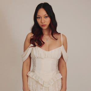 K&ouml;nnte beinhalten: Eine Frau in einem wei&szlig;en, schulterfreien Kleid mit einem Rock aus Spitze und einem taillierten Oberteil. Das Oberteil ist mit einem Muster aus kleinen wei&szlig;en Perlen verziert.