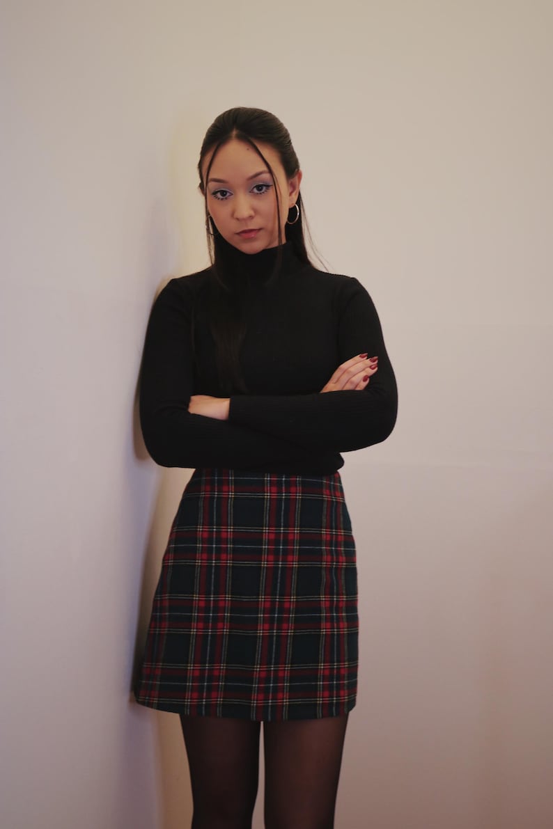 May include: A black turtleneck sweater paired with a plaid mini skirt in shades of green, red, and yellow. The outfit is completed with black tights. The model is leaning against a wall.