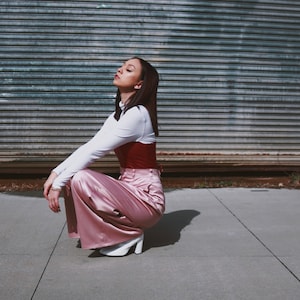 May include: A woman wearing a white long-sleeved shirt, a red corset, and pink satin pants. She is sitting on a concrete surface with a metal garage door in the background.