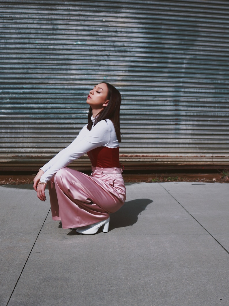 May include: A woman wearing a white long-sleeved shirt, a red corset, and pink satin pants. She is sitting on a concrete surface with a metal garage door in the background.
