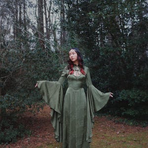 May include: A long, olive green dress with long, flared sleeves. The dress has a high neckline with a keyhole detail and a fitted bodice. The model is standing outdoors in a forest setting.