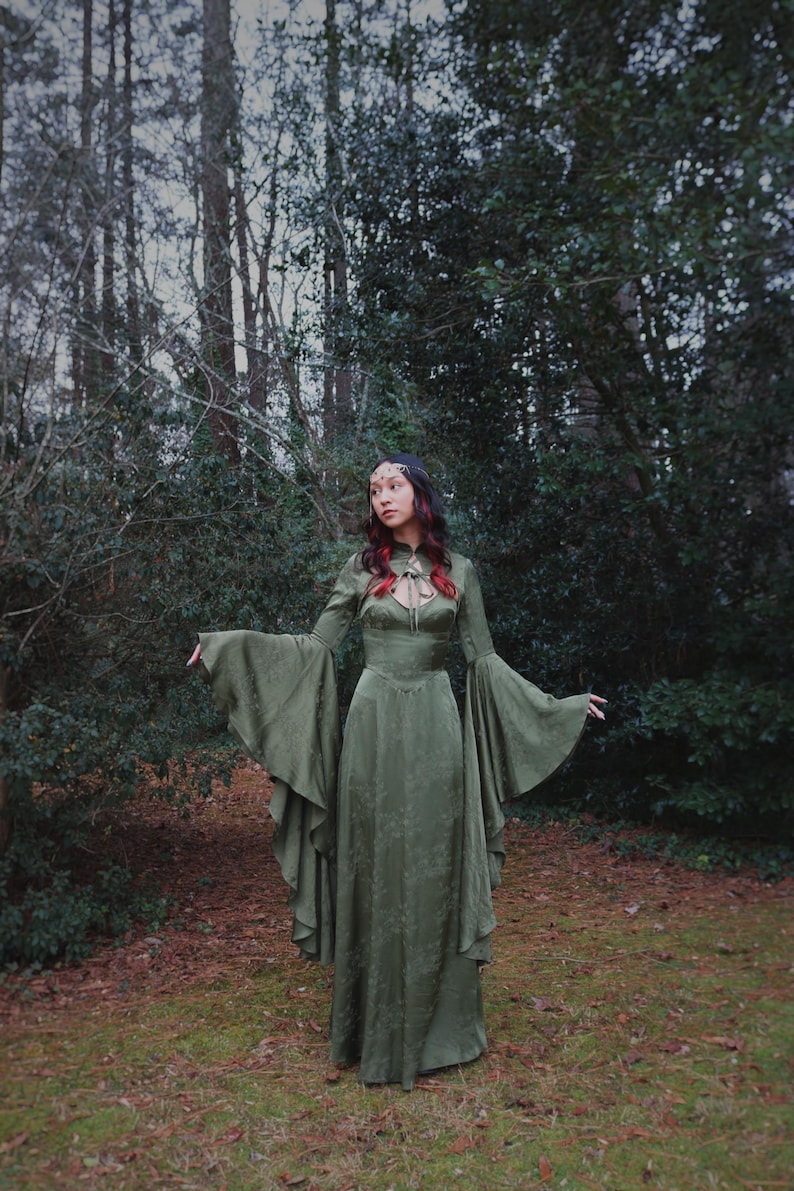 May include: A long, olive green dress with long, flared sleeves. The dress has a high neckline with a keyhole detail and a fitted bodice. The model is standing outdoors in a forest setting.