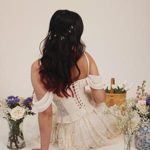 K&ouml;nnte beinhalten: Eine Frau in einem wei&szlig;en Spitzenkleid mit einem wei&szlig;en Korsett-Oberteil und einer Blumenkrone. Sie sitzt auf einem wei&szlig;en Hintergrund mit Blumen in Vasen.