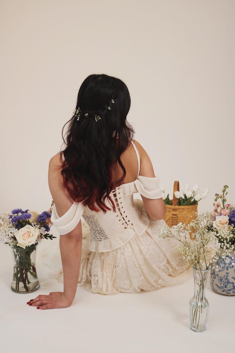 K&ouml;nnte beinhalten: Eine Frau in einem wei&szlig;en Spitzenkleid mit einem wei&szlig;en Korsett-Oberteil und einer Blumenkrone. Sie sitzt auf einem wei&szlig;en Hintergrund mit Blumen in Vasen.