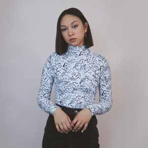 May include: A woman with short dark hair wearing a white long-sleeved shirt with a black and white abstract print and black jeans.