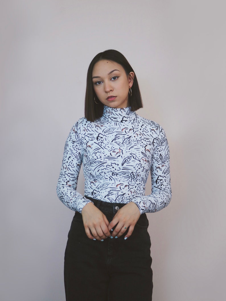 May include: A woman with short dark hair wearing a white long-sleeved shirt with a black and white abstract print and black jeans.