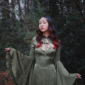 May include: An olive green dress with long, flared sleeves and a keyhole neckline. The dress features a floral pattern and is worn with a decorative headpiece. The person in the image has dark hair with red highlights, posing in a forest setting.