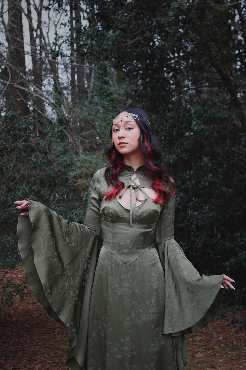 May include: An olive green dress with long, flared sleeves and a keyhole neckline. The dress features a floral pattern and is worn with a decorative headpiece. The person in the image has dark hair with red highlights, posing in a forest setting.