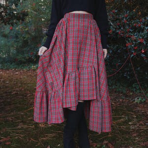 May include: A red and green plaid skirt with a tiered ruffle hem. The skirt is paired with a black long-sleeve top and black boots. The skirt's pattern is a classic tartan design, suitable for fall or winter wear.