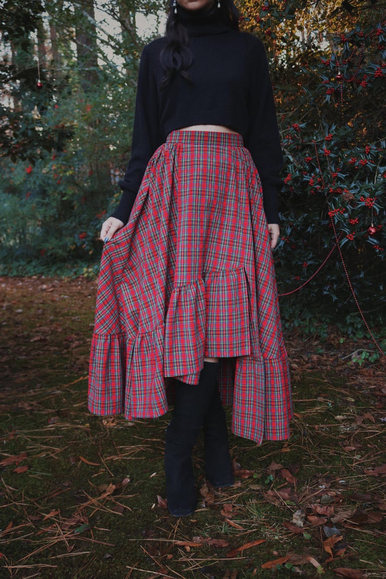 May include: A red and green plaid skirt with a tiered ruffle hem. The skirt is paired with a black long-sleeve top and black boots. The skirt's pattern is a classic tartan design, suitable for fall or winter wear.