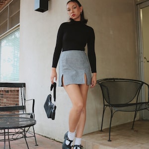 May include: A woman wearing a black turtleneck sweater, a gray and white plaid mini skirt with side slits, black and white saddle shoes, and white socks. She is holding a black handbag.