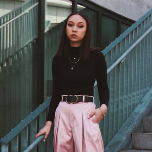 May include: A woman wearing a black mock neck shirt with a gold chain necklace and a black belt with a gold buckle. She is wearing pink wide-leg pants and has her hand in her pocket.