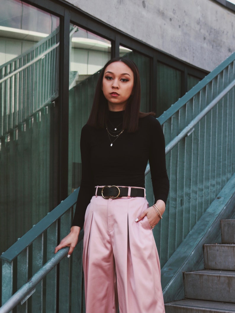 May include: A woman wearing a black mock neck shirt with a gold chain necklace and a black belt with a gold buckle. She is wearing pink wide-leg pants and has her hand in her pocket.