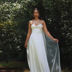 May include: A white wedding dress with a fitted bodice and flowing skirt. The dress features a criss-cross neckline and a sheer, flowing cape. The image is set outdoors with a backdrop of green foliage.