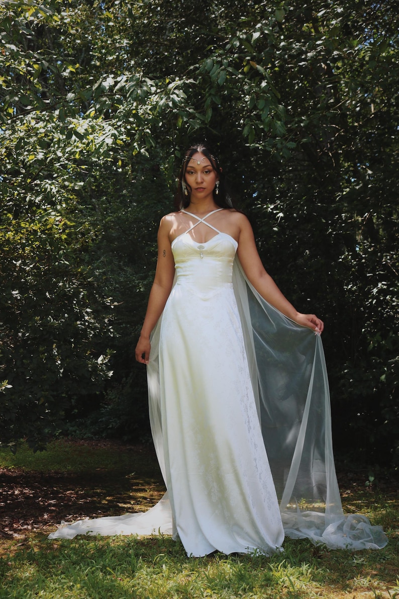 May include: A white wedding dress with a fitted bodice and flowing skirt. The dress features a criss-cross neckline and a sheer, flowing cape. The image is set outdoors with a backdrop of green foliage.