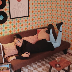 May include: A woman in a black outfit and light blue leggings reclines on a brown sofa. The background features a geometric patterned wallpaper and a framed abstract art piece. Vinyl records and a small wooden table with a drink are also visible.
