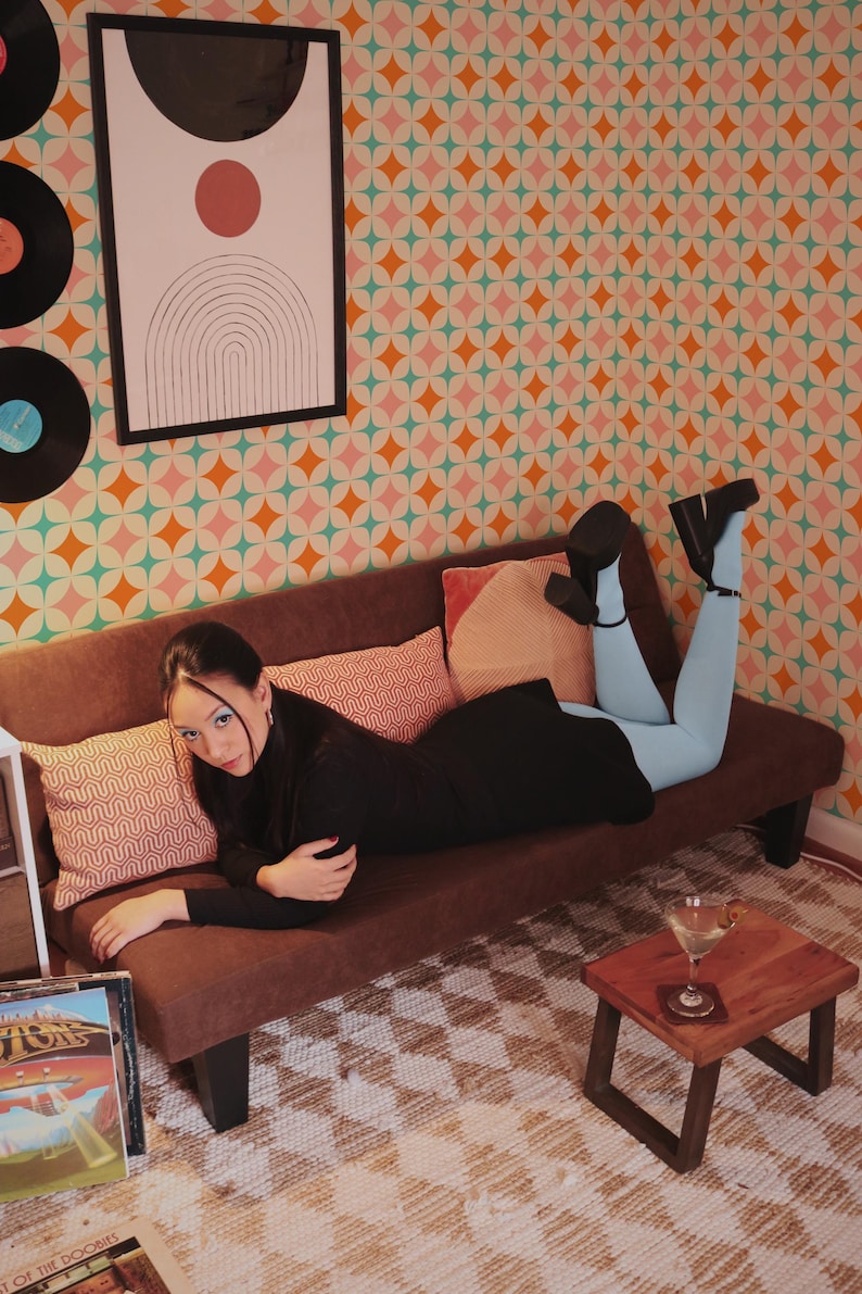 May include: A woman in a black outfit and light blue leggings reclines on a brown sofa. The background features a geometric patterned wallpaper and a framed abstract art piece. Vinyl records and a small wooden table with a drink are also visible.