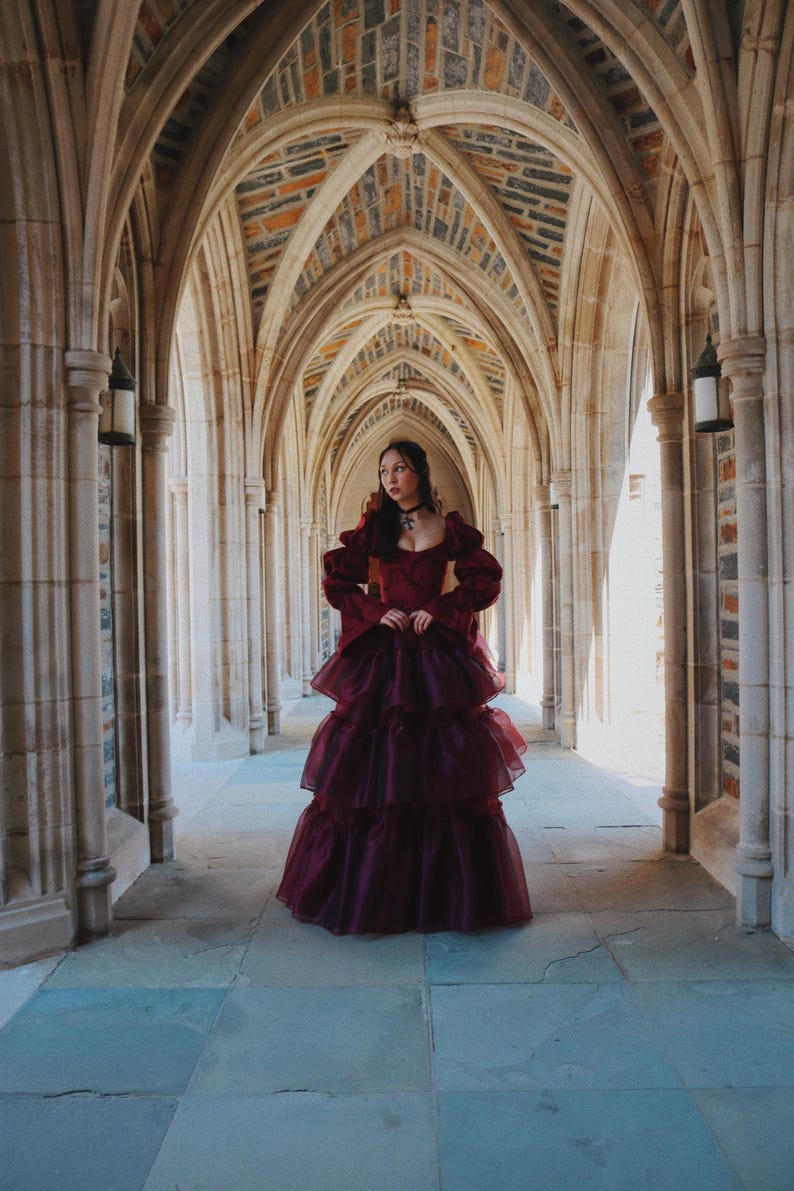 K&ouml;nnte beinhalten: Ein burgunderfarbenes, mehrschichtiges Abendkleid mit langen &Auml;rmeln und einem quadratischen Ausschnitt. Das Kleid wird in einem gew&ouml;lbten Steinflur mit einer Ziegeldecke getragen. Das Kleid hat einen geschichteten Rock und wird mit einem schwarzen Halsband erg&auml;nzt.