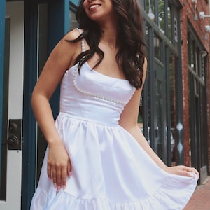 K&ouml;nnte beinhalten: Ein wei&szlig;es Satin-Kleid mit einem perlenbesetzten Riemendetail und einem R&uuml;schen-Saum. Das Kleid ist am Oberteil figurbetont und geht in einen Rock &uuml;ber.