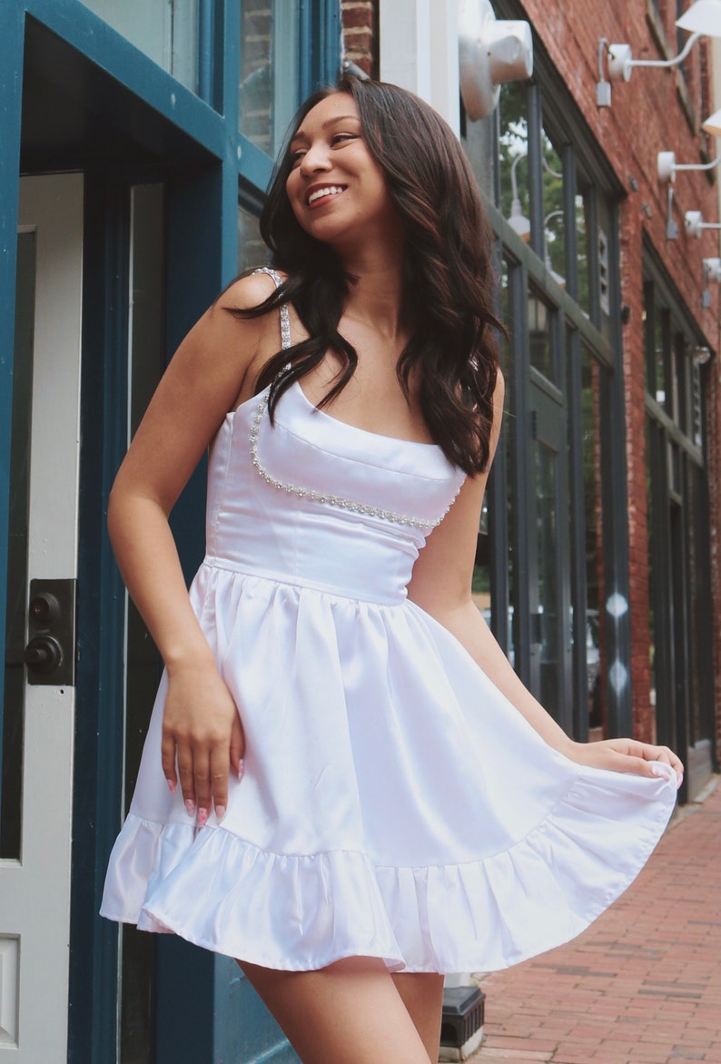 K&ouml;nnte beinhalten: Ein wei&szlig;es Satin-Kleid mit einem perlenbesetzten Riemendetail und einem R&uuml;schen-Saum. Das Kleid ist am Oberteil figurbetont und geht in einen Rock &uuml;ber.