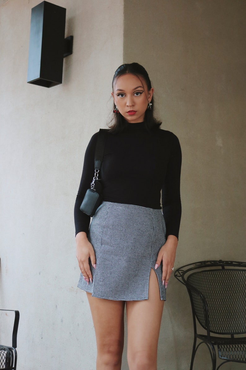 May include: A woman wearing a black mock neck shirt and a black and white checkered skirt with a side slit. She is wearing a small black crossbody bag with a silver chain.