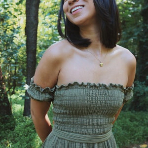 Op de afbeelding: Een vrouw die een groene off-shoulder jurk draagt met een gerimpelde halslijn en een bijpassende riem. Ze glimlacht en kijkt omhoog.