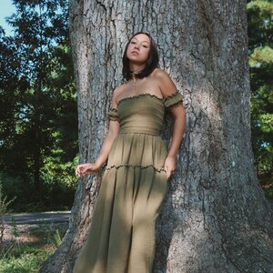 Op de afbeelding: Een vrouw in een groene off-shoulder jurk met een gelaagde rok staat voor een grote boom.