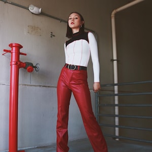 May include: A woman wearing a white and black long-sleeved shirt with a red leather belt and red leather pants. She is standing in front of a red fire hydrant and a metal gate.
