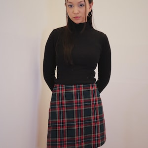May include: A black turtleneck sweater paired with a plaid mini skirt in green, red, and white. The skirt features a classic tartan pattern. The outfit is completed with black tights.