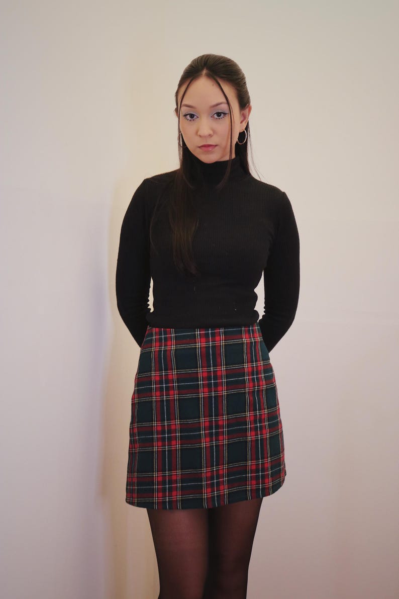May include: A black turtleneck sweater paired with a plaid mini skirt in green, red, and white. The skirt features a classic tartan pattern. The outfit is completed with black tights.
