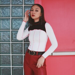 May include: A woman wearing a white long-sleeved shirt with black and pink trim, a gold belt, and red pants. She is standing in front of a wall with glass blocks.