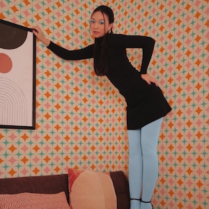 May include: A person wearing a black dress and light blue tights, standing on a brown sofa. The background features a colorful wallpaper with a geometric pattern. The person is leaning against a wall with a framed artwork.