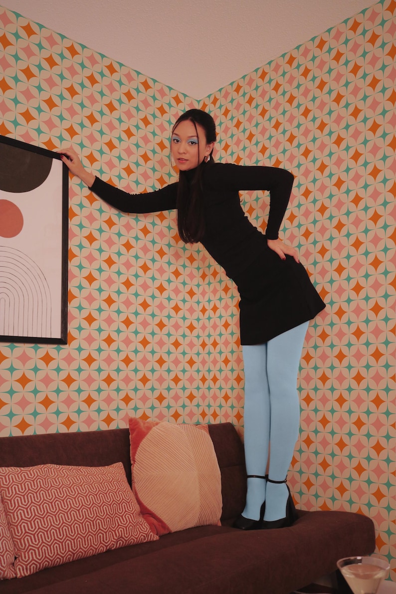 May include: A person wearing a black dress and light blue tights, standing on a brown sofa. The background features a colorful wallpaper with a geometric pattern. The person is leaning against a wall with a framed artwork.