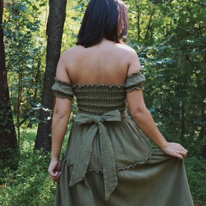 Op de afbeelding: Een vrouw draagt een groene, off-shoulder maxi-jurk met een gerimpelde zoom en een strik in de taille. De jurk is gemaakt van een lichtgewicht, vloeiende stof. Ze staat in een bosrijke omgeving.