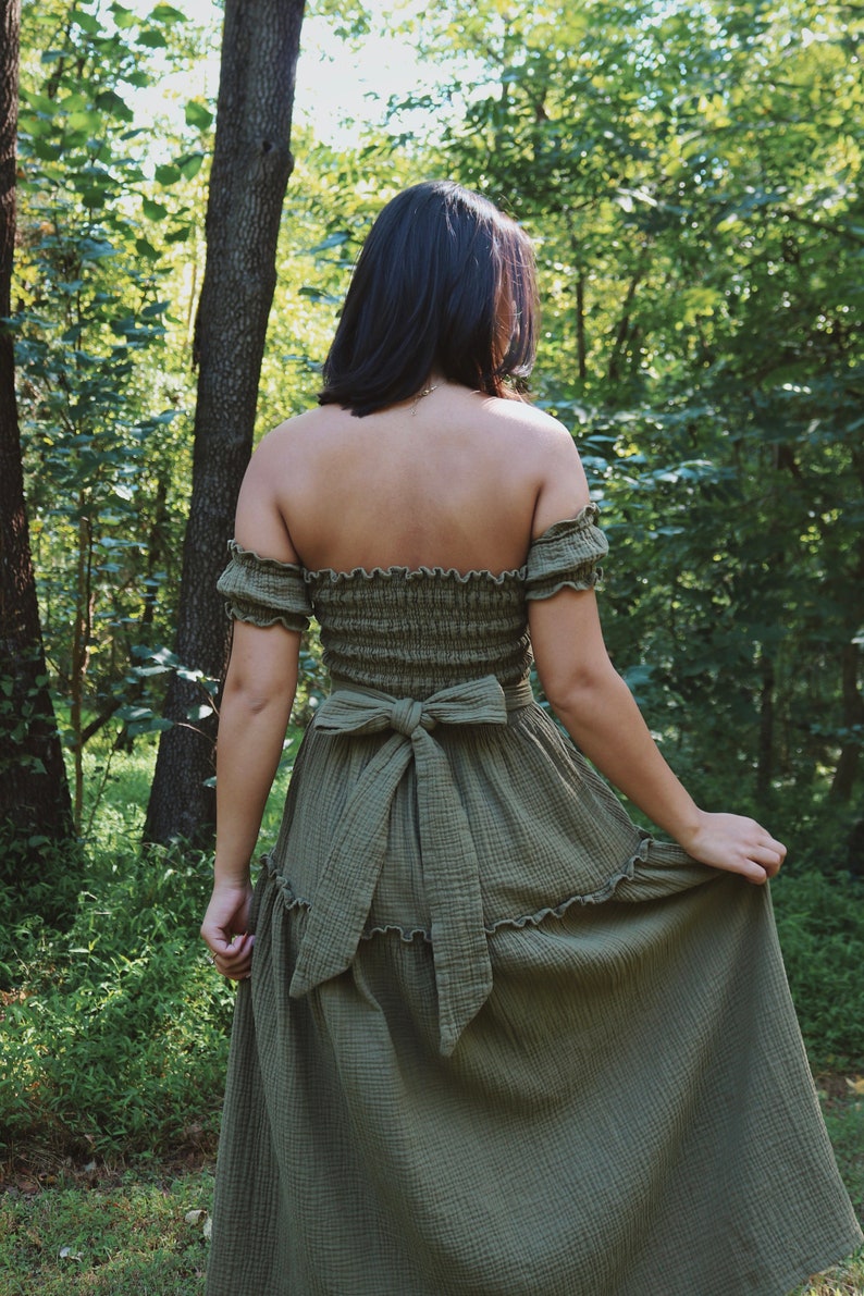 Op de afbeelding: Een vrouw draagt een groene, off-shoulder maxi-jurk met een gerimpelde zoom en een strik in de taille. De jurk is gemaakt van een lichtgewicht, vloeiende stof. Ze staat in een bosrijke omgeving.